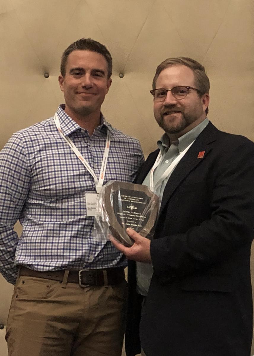Photo of Dustin Loy holding the award for excellence in diagnostic microbiology and standing next to the award presenter.