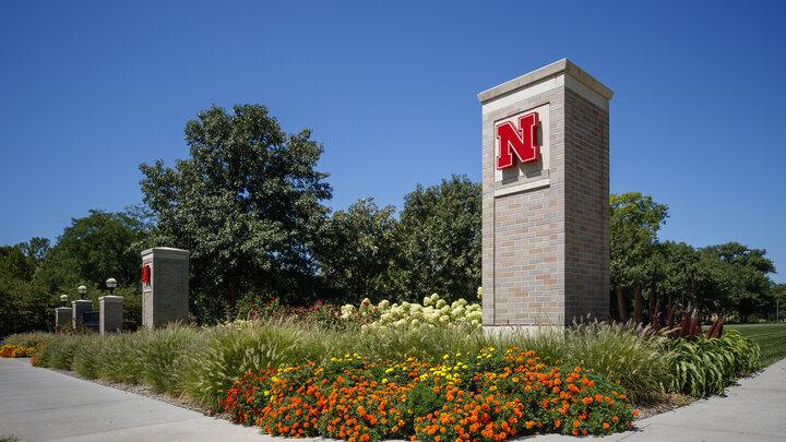 Two brick columns with red Husker Ns on the front are seen behind colorful flower beds on a clear day.