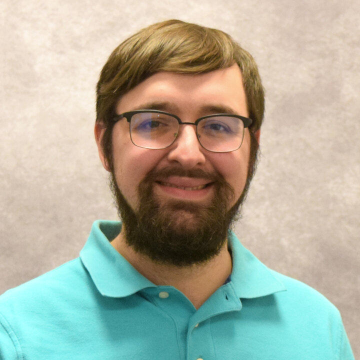 A studio portrait of Dr. Joel Di Bernardo. He is wearing a blue polo shirt and has glasses and a beard.
