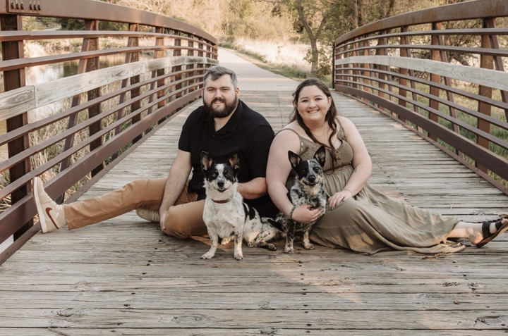 Jayden and her fiancé Christian sitting on a wooden bridge with two dogs, surrounded by greenery.