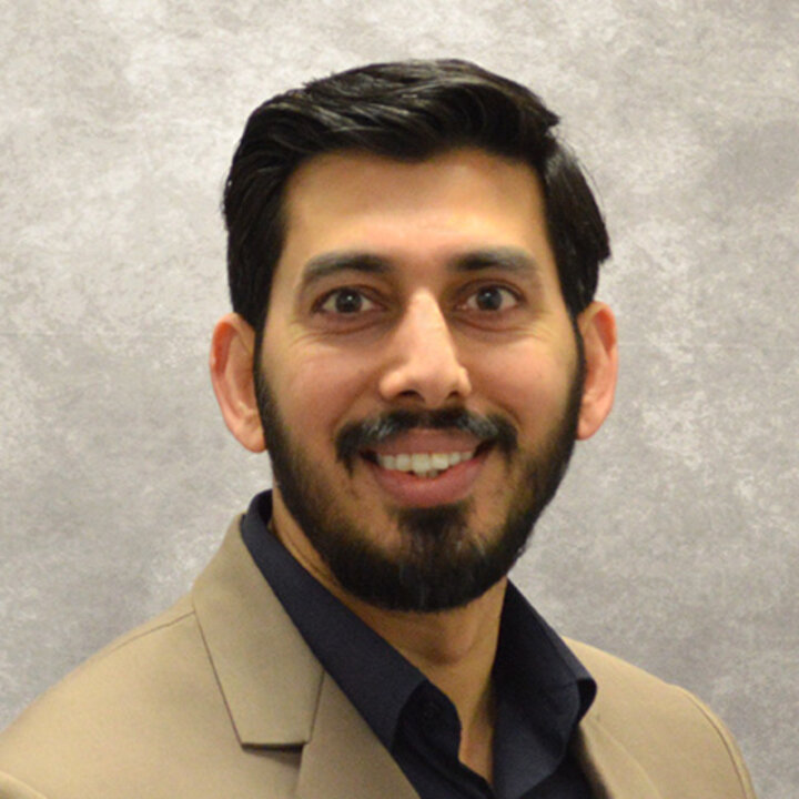Studio photo of Ahsan Naveed. Ahsan is wearing a tan jacket over a black button-up shirt. He has a beard.
