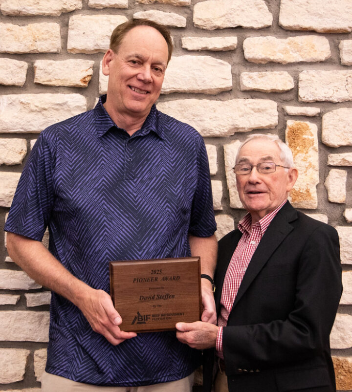 Dr. David Steffen stands in front of a stone wall. He is holding a plaque being presented to him by a representative from the Beef Improvement Federation